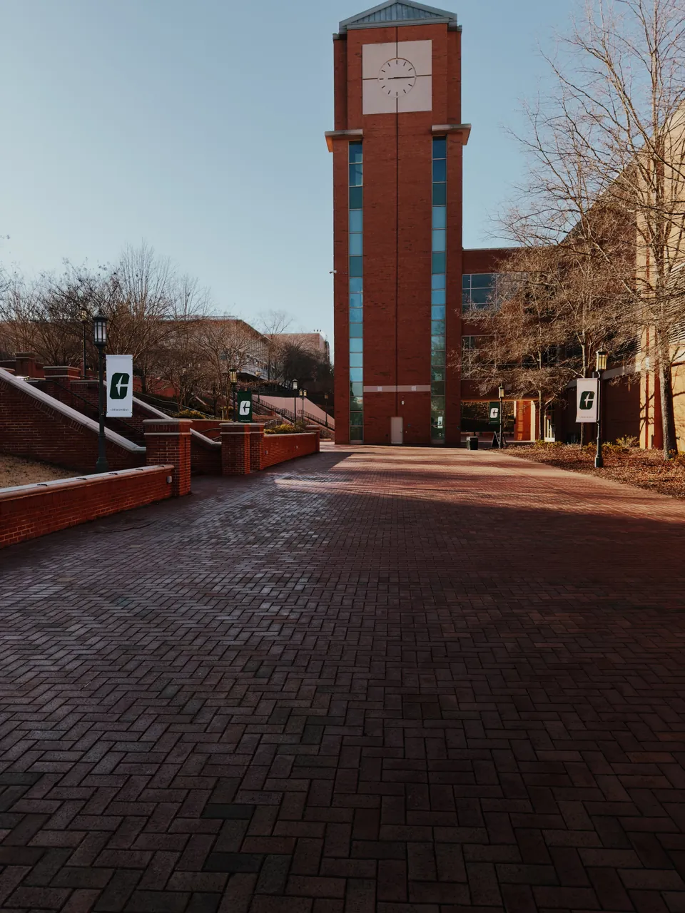 A brick clock tower at the end of an empty herringbone brick walkway, bare winter trees and green university banners either side.