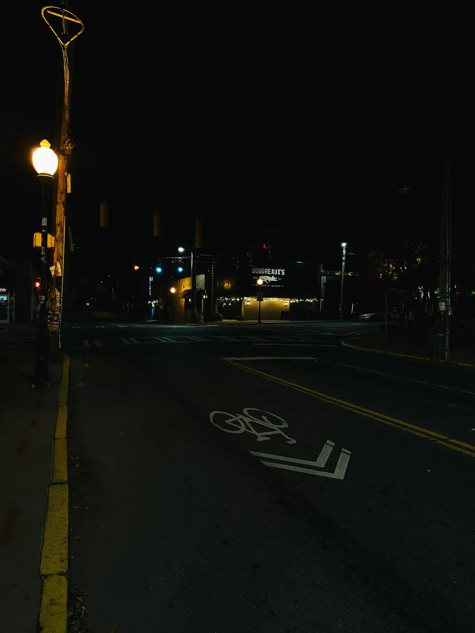An empty street at night, a single amber street lamp on the left, a painted bike lane arrow in the road and a lit bar sign in the distance.