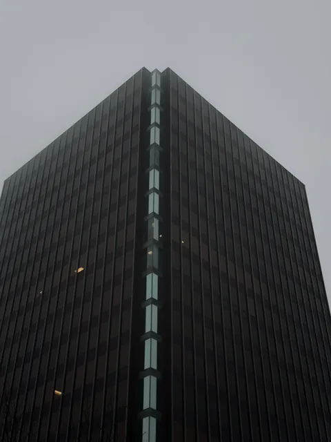Looking up at a dark glass office tower in fog, a single column of pale lit windows running up its corner to a flat grey sky.
